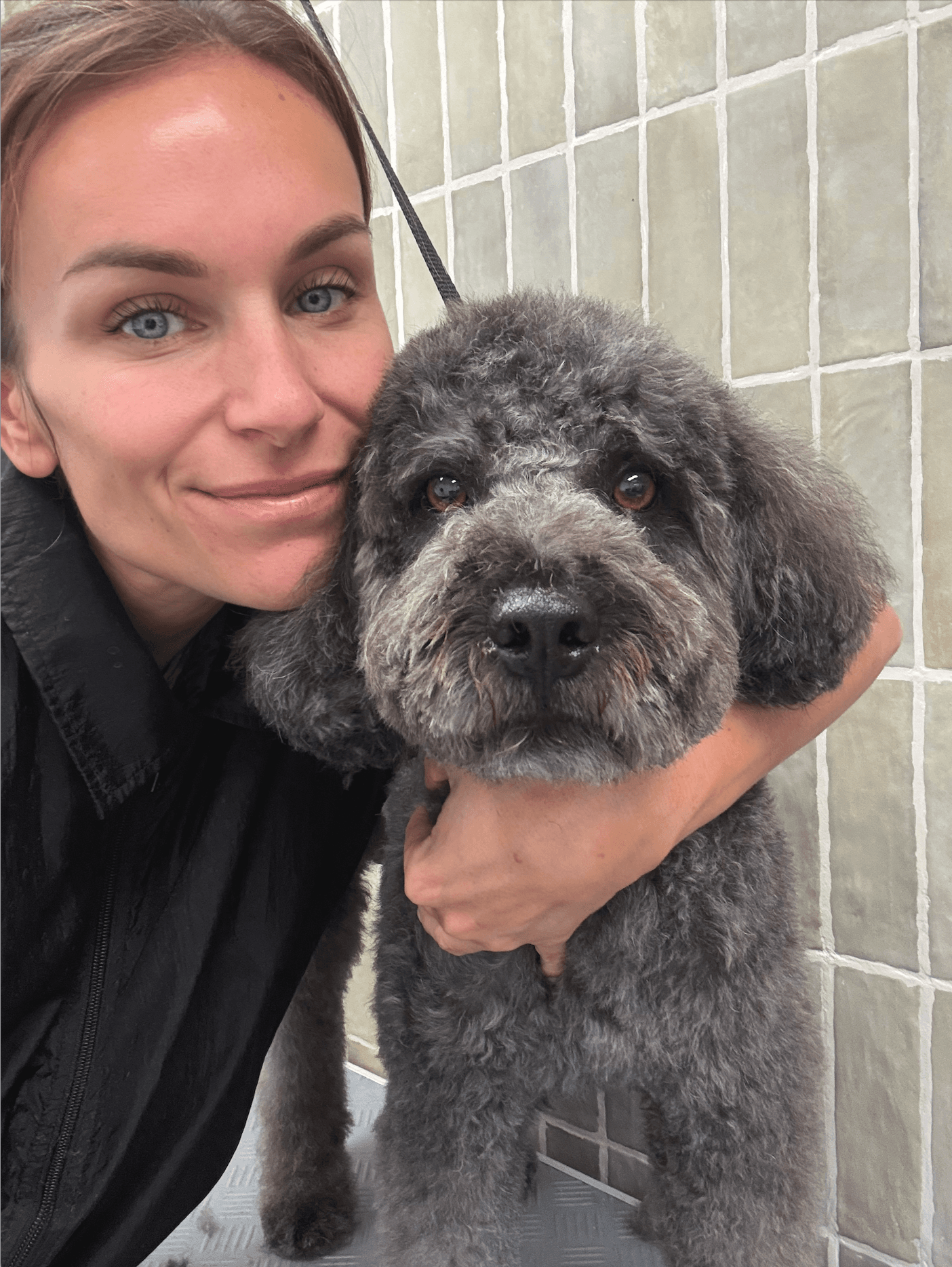 Une femme souriante aux yeux bleus pose avec un chien gris bouclé devant des carreaux.